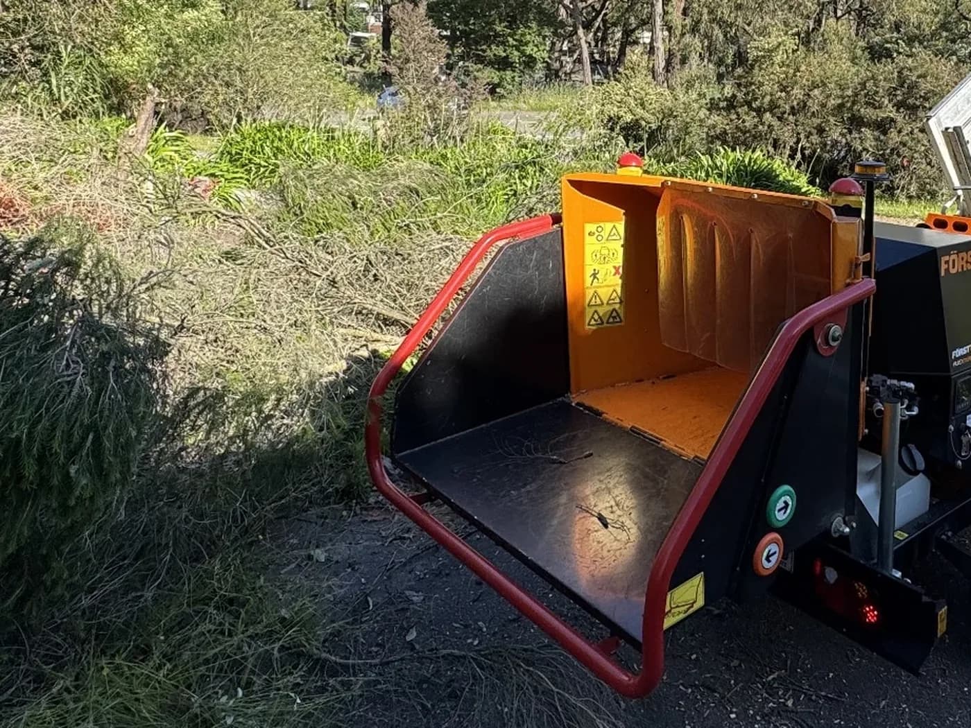 A wood chipper's open infeed hopper beside a pile of cut conifer branches on a driveway