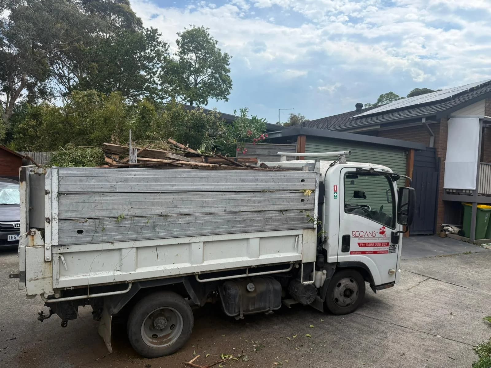 Regan's tipper truck, sign-written with the business name and phone number, loaded with timber and branches in a driveway