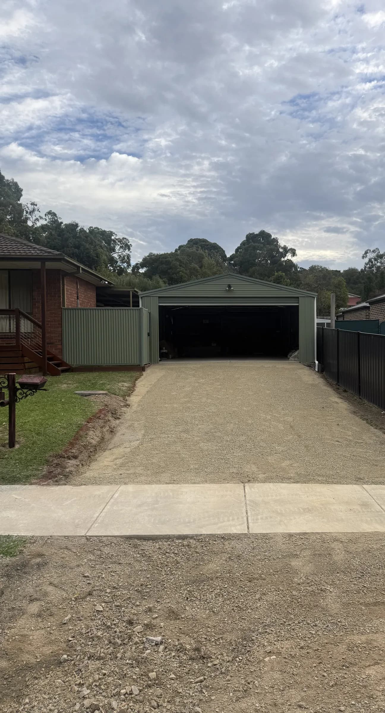 New crushed-rock driveway crossing a concrete footpath to a green steel shed