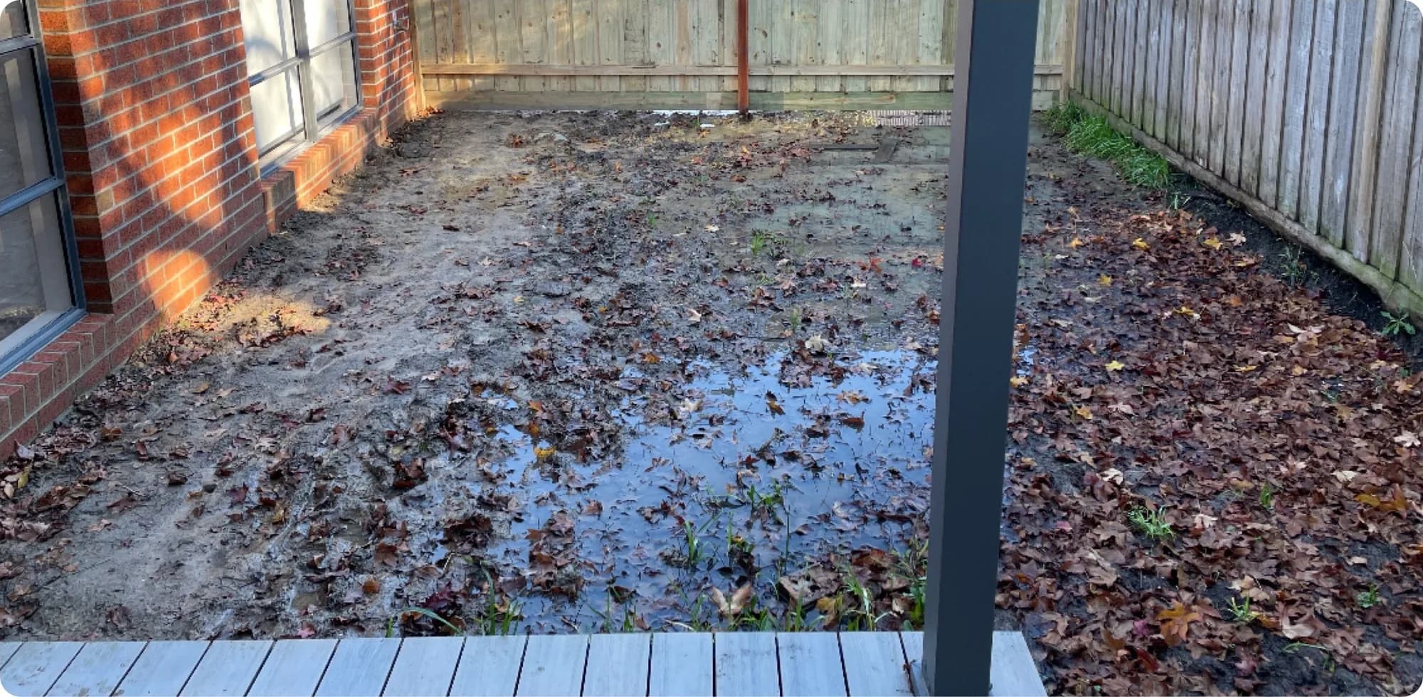 Narrow backyard of bare mud, standing water and fallen leaves beside a brick house
