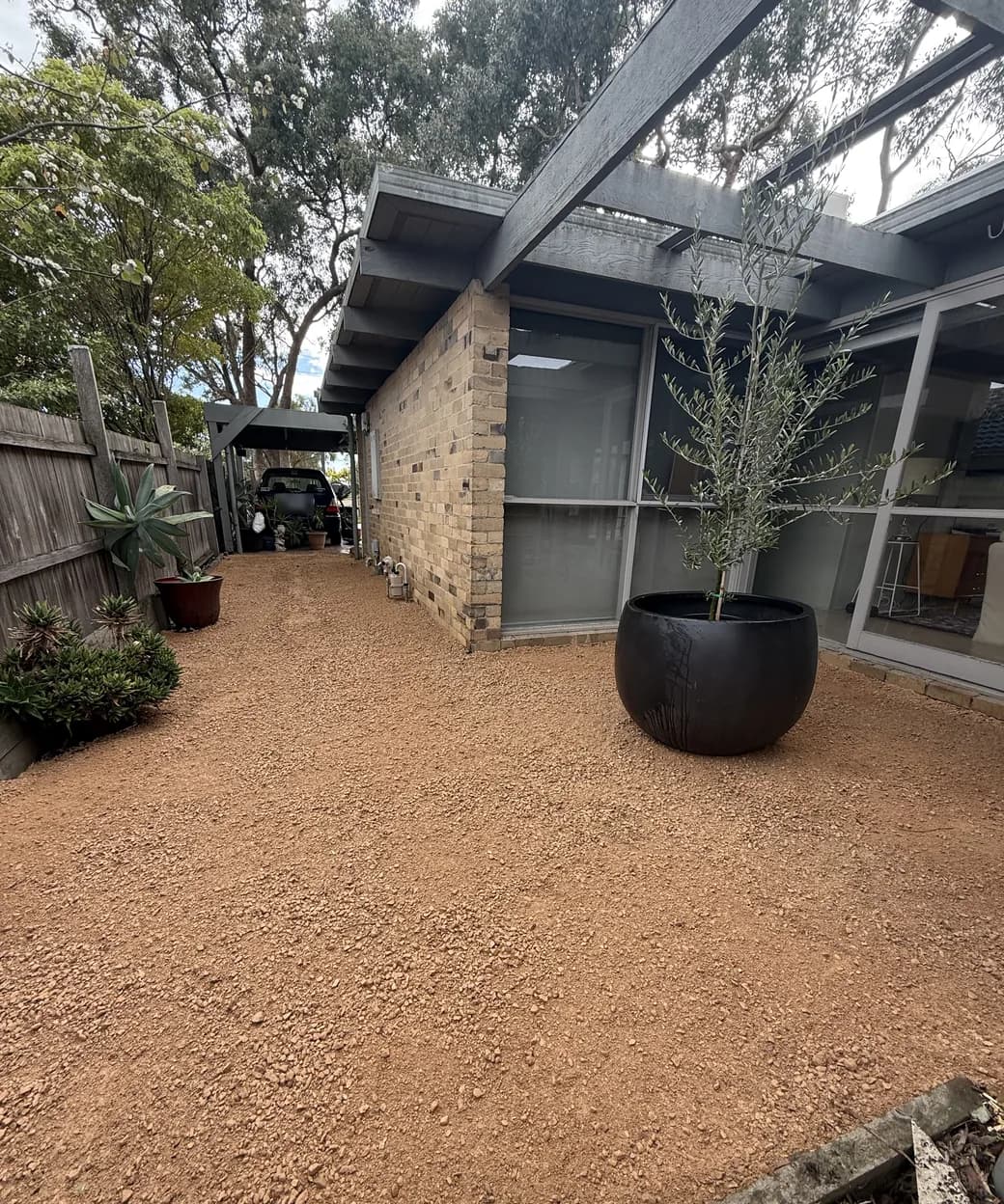 A courtyard laid with compacted crushed rock beside a brick house, with a potted olive tree